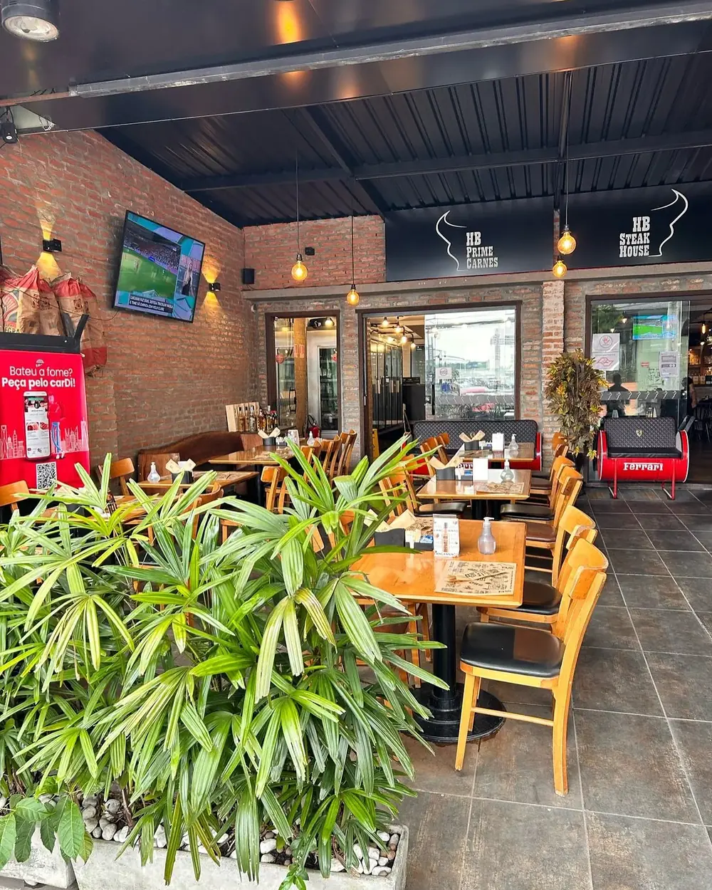 Salão da HB SteakHouse com mesas de madeira, cadeiras amarelas e parede de tijolo